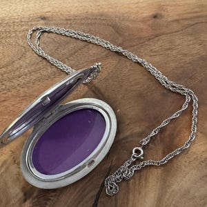 Puede incluir: Collar con colgante ovalado plateado con tapa abierta que revela un interior p&uacute;rpura. El relicario est&aacute; sujeto a una delicada cadena, sobre una superficie de madera. Una pieza de joyer&iacute;a cl&aacute;sica.