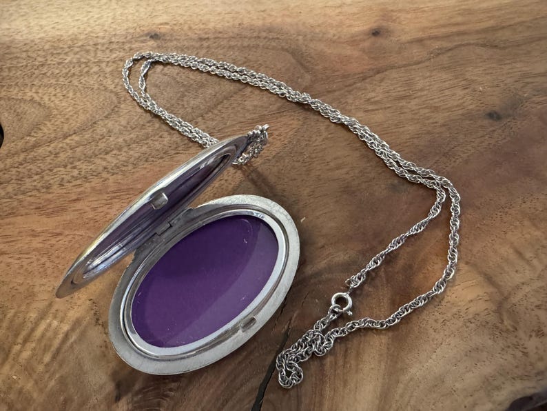 Puede incluir: Collar con colgante ovalado plateado con tapa abierta que revela un interior p&uacute;rpura. El relicario est&aacute; sujeto a una delicada cadena, sobre una superficie de madera. Una pieza de joyer&iacute;a cl&aacute;sica.