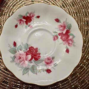 May include: A white porcelain saucer with a scalloped edge features a delicate floral design of pink and red roses with green leaves. The saucer is perfect for serving tea or coffee.