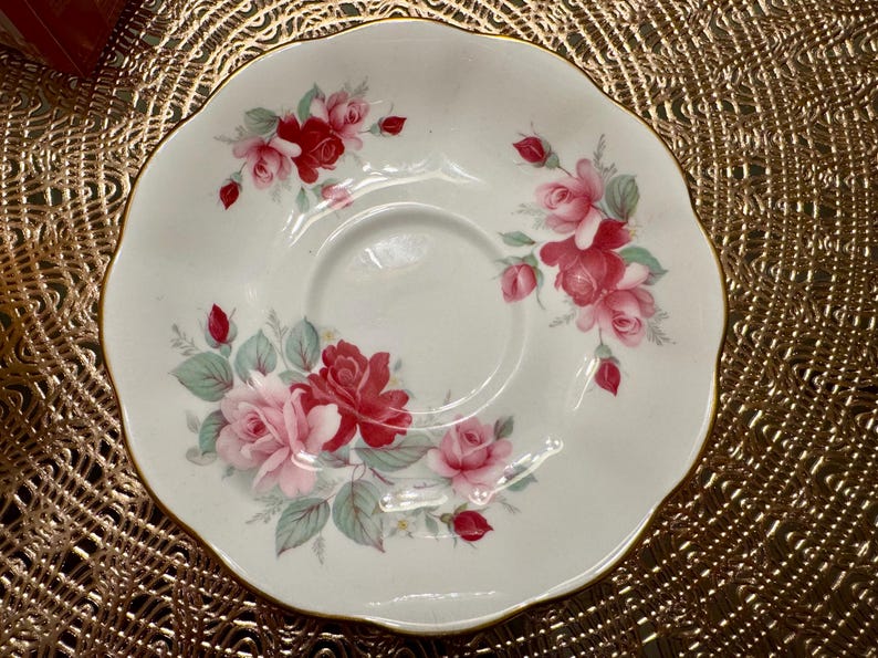 May include: A white porcelain saucer with a scalloped edge features a delicate floral design of pink and red roses with green leaves. The saucer is perfect for serving tea or coffee.