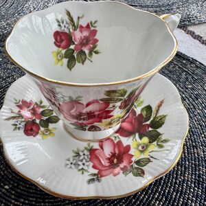 Puede incluir: Un juego de taza y platillo de t&eacute; blanco con un patr&oacute;n floral con rosas rosas, rojas y amarillas. La taza y el platillo tienen un borde dorado.