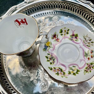 May include: A white teacup and saucer set with floral and ribbon designs, and gold trim. The teacup features a Canadian flag. The set is displayed on a silver serving tray, perfect for tea parties or collectors.