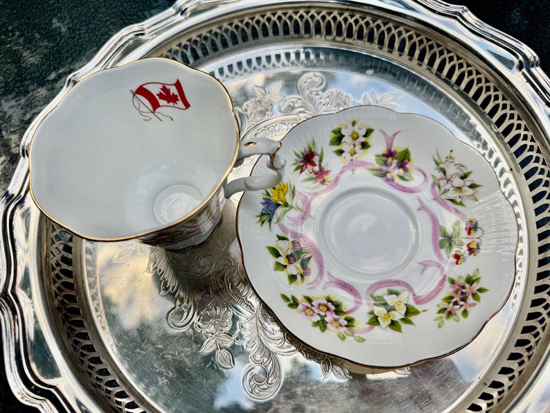 May include: A white teacup and saucer set with floral and ribbon designs, and gold trim. The teacup features a Canadian flag. The set is displayed on a silver serving tray, perfect for tea parties or collectors.