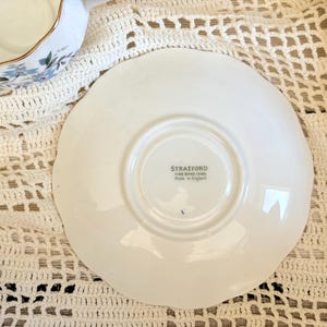 May include: A white ceramic saucer with a scalloped edge, featuring a central stamp that reads STRATFORD FINE BONE CHINA Made in England. The saucer is circular with a raised center ring and a smooth, glossy finish on its surface.