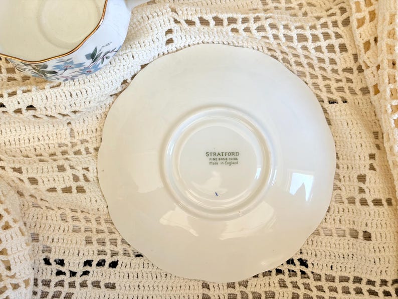 May include: A white ceramic saucer with a scalloped edge, featuring a central stamp that reads STRATFORD FINE BONE CHINA Made in England. The saucer is circular with a raised center ring and a smooth, glossy finish on its surface.