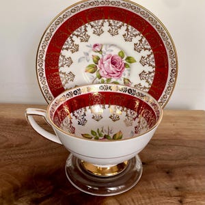 Pode incluir: Conjunto de ch&aacute; branco com um design vermelho e dourado ornamentado. A ch&aacute;vena apresenta um padr&atilde;o floral de rosa rosa e acabamentos dourados. O pires tem um design a condizer. O conjunto est&aacute; sobre um suporte de vidro transparente.