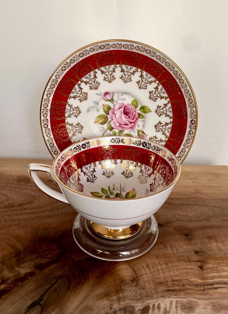 Pode incluir: Conjunto de ch&aacute; branco com um design vermelho e dourado ornamentado. A ch&aacute;vena apresenta um padr&atilde;o floral de rosa rosa e acabamentos dourados. O pires tem um design a condizer. O conjunto est&aacute; sobre um suporte de vidro transparente.