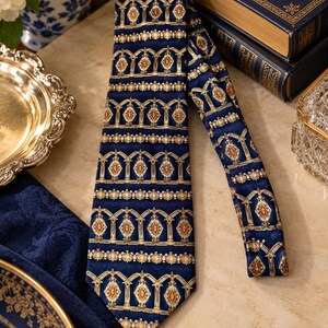 May include: A navy blue necktie featuring a repeating pattern of gold and orange architectural arch motifs arranged in horizontal bands. The tie has a pointed tip and a smooth, satin-like finish with intricate detailing throughout the fabric design.