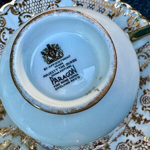 May include: A white fine bone china teacup and saucer set featuring an intricate gold filigree pattern. The base of the cup displays a black stamp reading BY APPOINTMENT TO HER MAJESTY THE QUEEN CHINA POTTERS PARAGON FINE BONE CHINA ENGLAND REG'D.