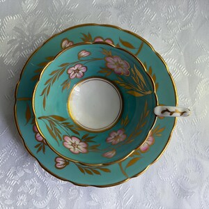 May include: A turquoise teacup and saucer set featuring a white interior, scalloped edges, and gold trim. The exterior of the cup and the saucer are decorated with hand-painted pink flowers and gold leaf foliage patterns throughout the surface.