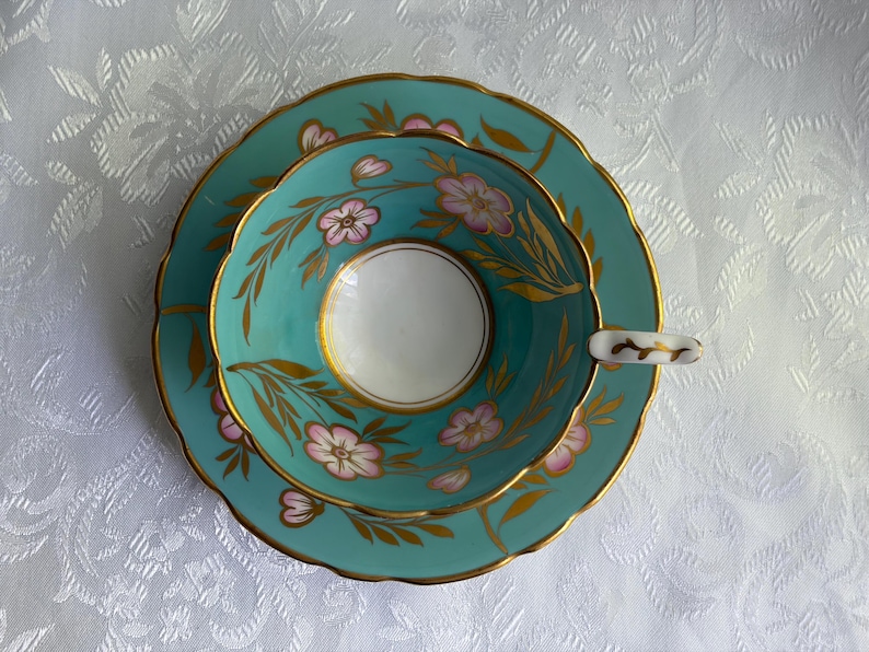 May include: A turquoise teacup and saucer set featuring a white interior, scalloped edges, and gold trim. The exterior of the cup and the saucer are decorated with hand-painted pink flowers and gold leaf foliage patterns throughout the surface.