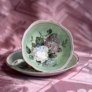 May include: A porcelain teacup and saucer set featuring a pale mint green interior and exterior. The cup is decorated with a floral pattern of white, blue, and purple hydrangea blossoms and green leaves, finished with a delicate gold trim on the rim.