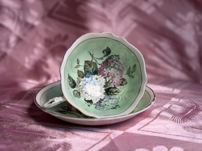 May include: A porcelain teacup and saucer set featuring a pale mint green interior and exterior. The cup is decorated with a floral pattern of white, blue, and purple hydrangea blossoms and green leaves, finished with a delicate gold trim on the rim.