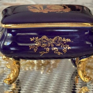 May include: A navy blue porcelain trinket box with gold accents. The box features a gold bow and floral design on the front and a gold design on the lid. The box has gold-colored feet.