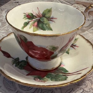May include: A vintage teacup and saucer set. The white porcelain cup and saucer feature a red rose and green leaf design. The rims are trimmed with gold. The cup has a delicate handle.