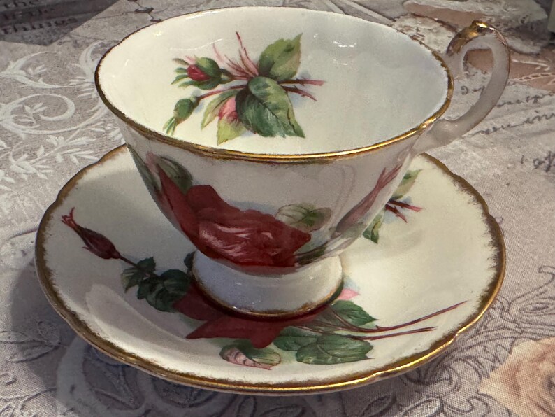 May include: A vintage teacup and saucer set. The white porcelain cup and saucer feature a red rose and green leaf design. The rims are trimmed with gold. The cup has a delicate handle.