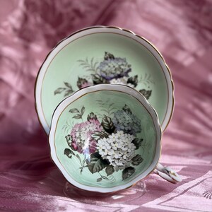 May include: A porcelain teacup and saucer set featuring a pale mint green background with a delicate floral pattern of white, pink, and blue hydrangea clusters. The edges of both the cup and saucer are scalloped and trimmed with a thin gold rim.