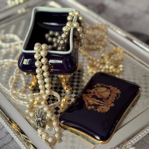 May include: A navy blue jewelry box with gold accents and a matching case with a gold illustration sit on a silver tray. Pearl necklaces and bracelets are draped around the box and tray, creating an elegant display.