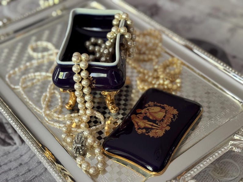 May include: A navy blue jewelry box with gold accents and a matching case with a gold illustration sit on a silver tray. Pearl necklaces and bracelets are draped around the box and tray, creating an elegant display.