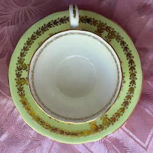May include: A pale green teacup and saucer set featuring a white interior and a gold-toned laurel leaf pattern along the rim of the saucer and the inner edge of the cup. The cup has a gold-trimmed handle and a scalloped edge on the saucer.