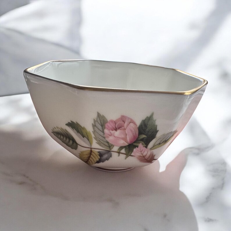 Pode incluir: Uma tigela octogonal creme com borda dourada e design floral. A tigela apresenta uma rosa rosa com folhas verdes e est&aacute; sobre um fundo branco.
