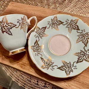 May include: A light blue teacup and saucer set with a gold leaf design. The teacup has a handle and the saucer has a scalloped edge.