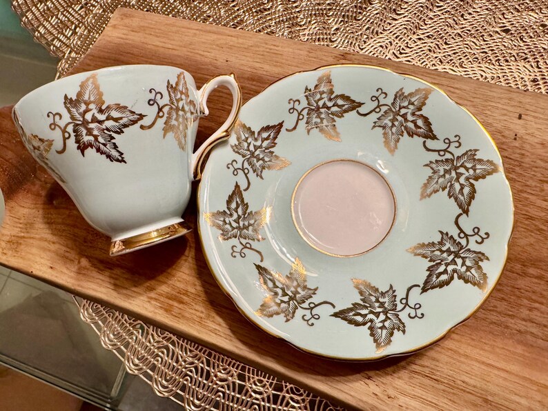 May include: A light blue teacup and saucer set with a gold leaf design. The teacup has a handle and the saucer has a scalloped edge.