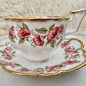 Puede incluir: Juego de taza y platillo blancos con dise&ntilde;o floral. La taza y el platillo est&aacute;n decorados con flores rosas y hojas verdes. Los bordes est&aacute;n adornados con oro. La taza tiene un asa angular.