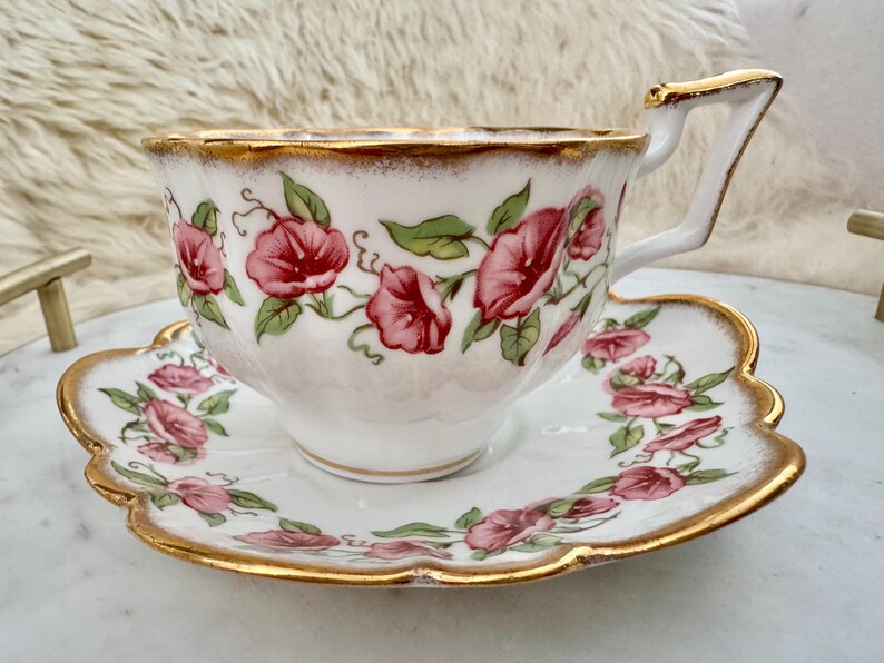 Puede incluir: Juego de taza y platillo blancos con dise&ntilde;o floral. La taza y el platillo est&aacute;n decorados con flores rosas y hojas verdes. Los bordes est&aacute;n adornados con oro. La taza tiene un asa angular.