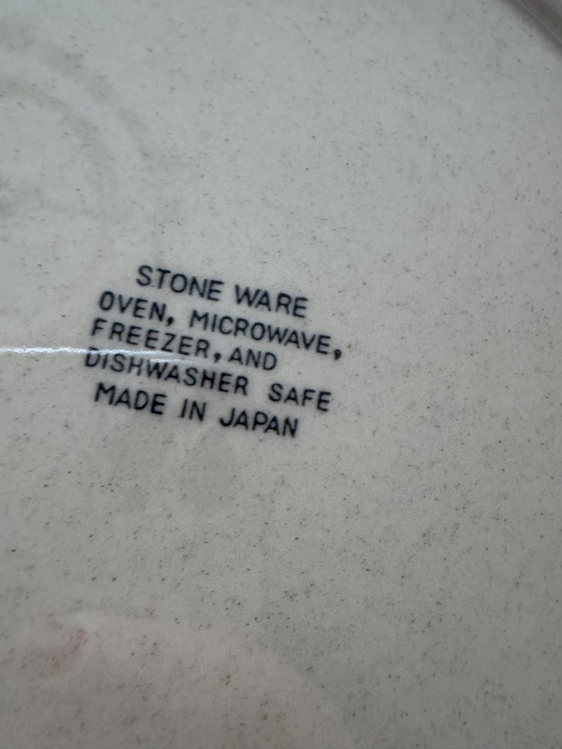 May include: Close-up of a speckled, off-white stoneware surface. Text in black reads: STONE WARE, OVEN, MICROWAVE, FREEZER, AND DISHWASHER SAFE, MADE IN JAPAN. The surface has a slightly textured appearance.