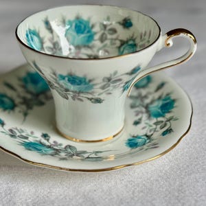 Peut inclure: Une tasse et une soucoupe blanches avec un motif floral de roses turquoise et de feuilles grises. La tasse et la soucoupe ont une bordure dor&eacute;e. La tasse a une anse incurv&eacute;e. L'ensemble est sur une surface blanche.