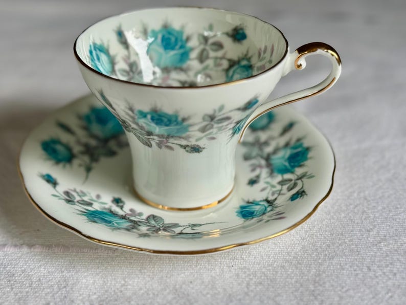 Peut inclure: Une tasse et une soucoupe blanches avec un motif floral de roses turquoise et de feuilles grises. La tasse et la soucoupe ont une bordure dor&eacute;e. La tasse a une anse incurv&eacute;e. L'ensemble est sur une surface blanche.
