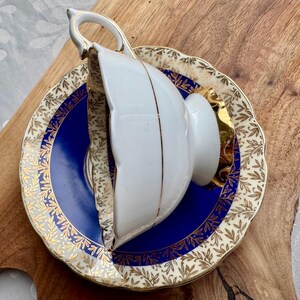 May include: A white teacup and saucer with gold trim and a blue and gold floral pattern. The saucer has a scalloped edge.