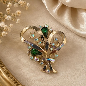 May include: Gold-toned vintage brooch with a floral design. The brooch features a central green teardrop-shaped stone, surrounded by smaller iridescent and clear crystals. The brooch has a ribbon-like design.