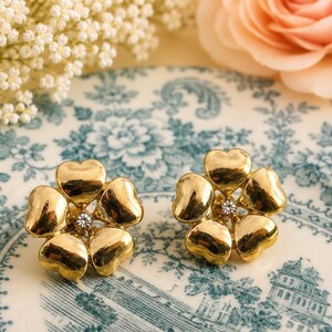 May include: A pair of gold-toned flower-shaped earrings. Each earring features five heart-shaped petals surrounding a small, clear crystal. The earrings are displayed on a vintage-style plate with a blue and white floral pattern.