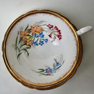 May include: A white teacup with a gold rim and a floral design featuring yellow, orange, red, blue, and green flowers. The cup has a scalloped edge.
