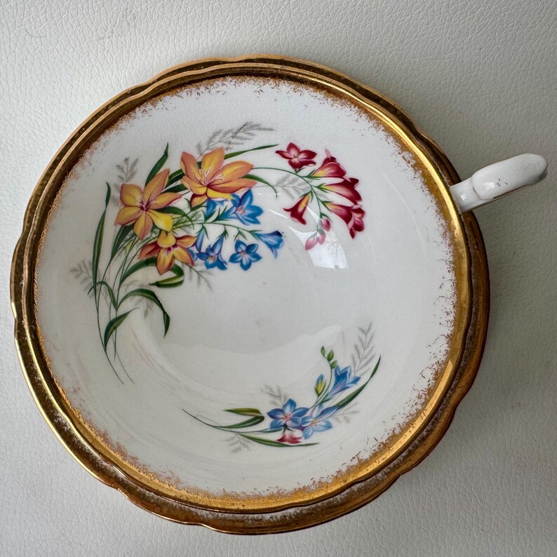 May include: A white teacup with a gold rim and a floral design featuring yellow, orange, red, blue, and green flowers. The cup has a scalloped edge.
