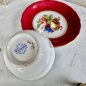 May include: A white bone china teacup and saucer with a red rim. The saucer has a floral design with fruit and a gold trim. The teacup has the words "Made in England, Bone Windsor China" printed on the bottom.