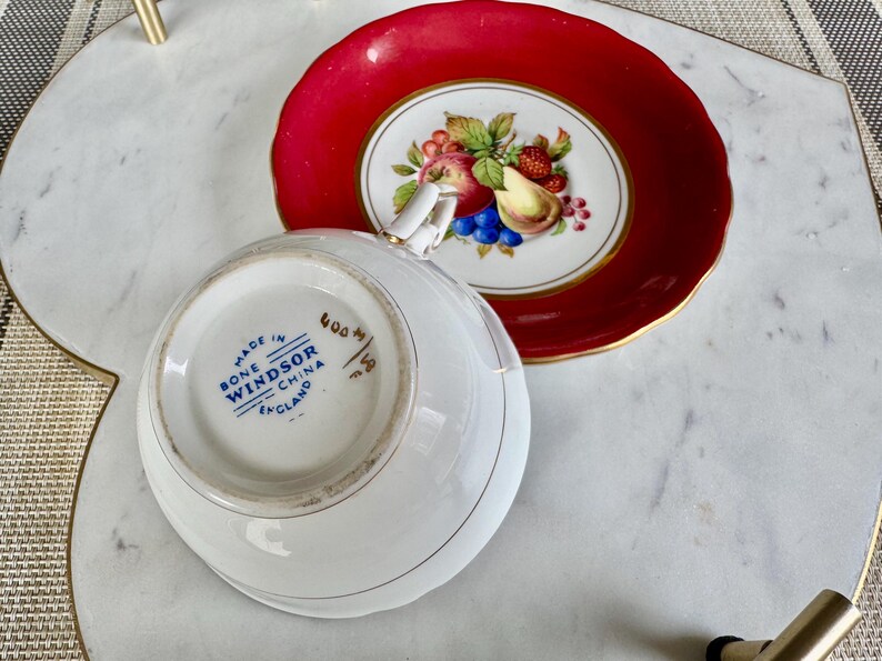 May include: A white bone china teacup and saucer with a red rim. The saucer has a floral design with fruit and a gold trim. The teacup has the words "Made in England, Bone Windsor China" printed on the bottom.