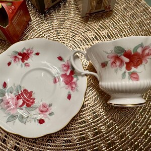May include: A white teacup and saucer set with a floral design featuring red, pink, and white roses. The set has a gold trim around the edges.