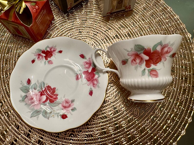 May include: A white teacup and saucer set with a floral design featuring red, pink, and white roses. The set has a gold trim around the edges.