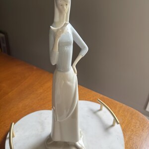 K&ouml;nnte beinhalten: Porzellanfigur einer Frau, die einen Vogel h&auml;lt. Die Figur tr&auml;gt ein langes, hellblaues Kleid mit langen &Auml;rmeln und einen wei&szlig;en Rock. Die Figur steht auf einem herzf&ouml;rmigen Marmortablett mit goldenen Griffen. Die Figur ist etwa 30 cm hoch.