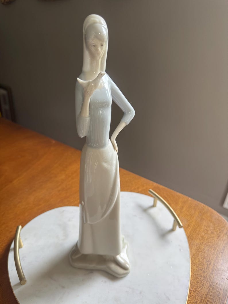 K&ouml;nnte beinhalten: Porzellanfigur einer Frau, die einen Vogel h&auml;lt. Die Figur tr&auml;gt ein langes, hellblaues Kleid mit langen &Auml;rmeln und einen wei&szlig;en Rock. Die Figur steht auf einem herzf&ouml;rmigen Marmortablett mit goldenen Griffen. Die Figur ist etwa 30 cm hoch.