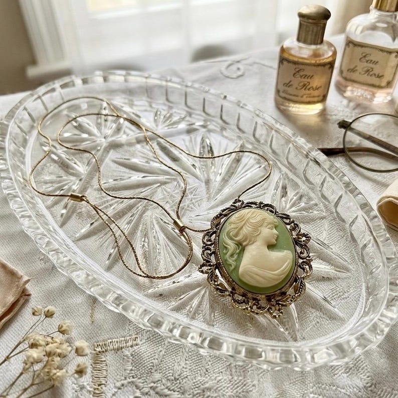 May include: A gold-toned necklace with a cameo pendant featuring a woman's profile in cream against a green background. The pendant is set in an ornate, antique-style frame. The necklace rests on a clear, oval-shaped glass tray.