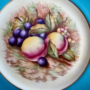May include: A decorative plate with a painted still life of fruit. The design features purple grapes, peaches with pink and yellow hues, and green leaves. The plate has a cream-colored center with a gold rim and a bright blue border.
