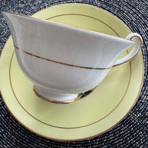 May include: A white teacup and saucer set with a yellow rim and gold trim. The teacup has a delicate handle and a rounded shape.