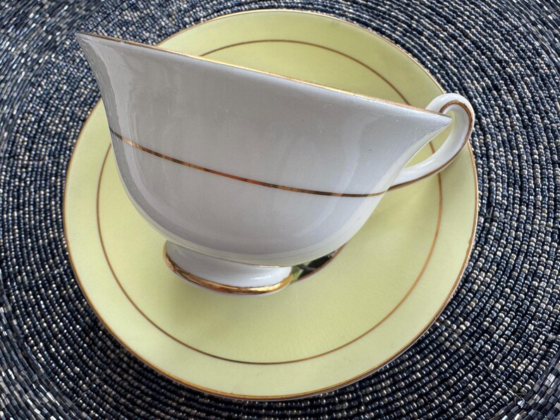 May include: A white teacup and saucer set with a yellow rim and gold trim. The teacup has a delicate handle and a rounded shape.