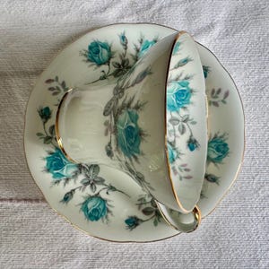 Peut inclure: Un service &agrave; th&eacute; vintage compos&eacute; d'une tasse et d'une soucoupe. La tasse et la soucoupe en porcelaine blanche sont orn&eacute;es d'un motif floral de roses turquoise et d'une bordure dor&eacute;e. La tasse est inclin&eacute;e, r&eacute;v&eacute;lant l'int&eacute;rieur et l'anse.