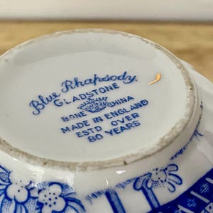 May include: Close-up of a white teacup's interior with blue floral and geometric patterns. The text "Blue Rhapsody Gladstone Bone China Made in England Estd Over 80 Years" is printed in blue.