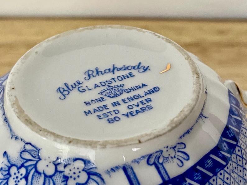 May include: Close-up of a white teacup's interior with blue floral and geometric patterns. The text "Blue Rhapsody Gladstone Bone China Made in England Estd Over 80 Years" is printed in blue.
